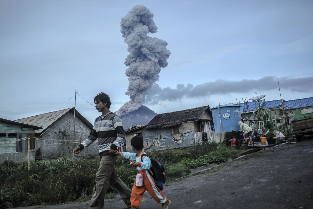 Warga melintas dengan latar belakang Gunung Sinabung mengeluarkan material vulkanik ketika erupsi, di Karo, Sumatera Utara, Sabtu (3/9). Aktivitas Gunung Sinabung berstatus Awas (level IV) sejak seminggu terakhir semakin meningkat ditandai dengan erupsi. ANTARA FOTO/Zulkarnain Ginting/im/foc/16.